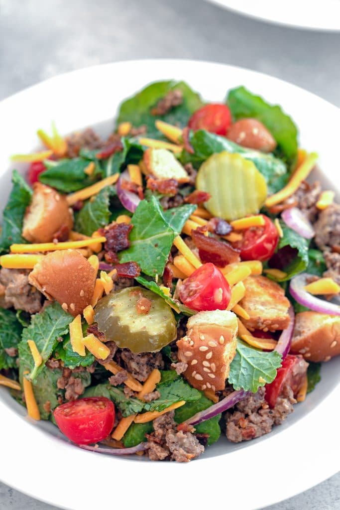 Overhead close-up view of bacon cheeseburger kale salad including baby kale, ground beef, shredded cheddar, dill pickle rounds, chopped tomatoes, red onion, crumbled bacon, and hamburger bun croutons in a white bowl