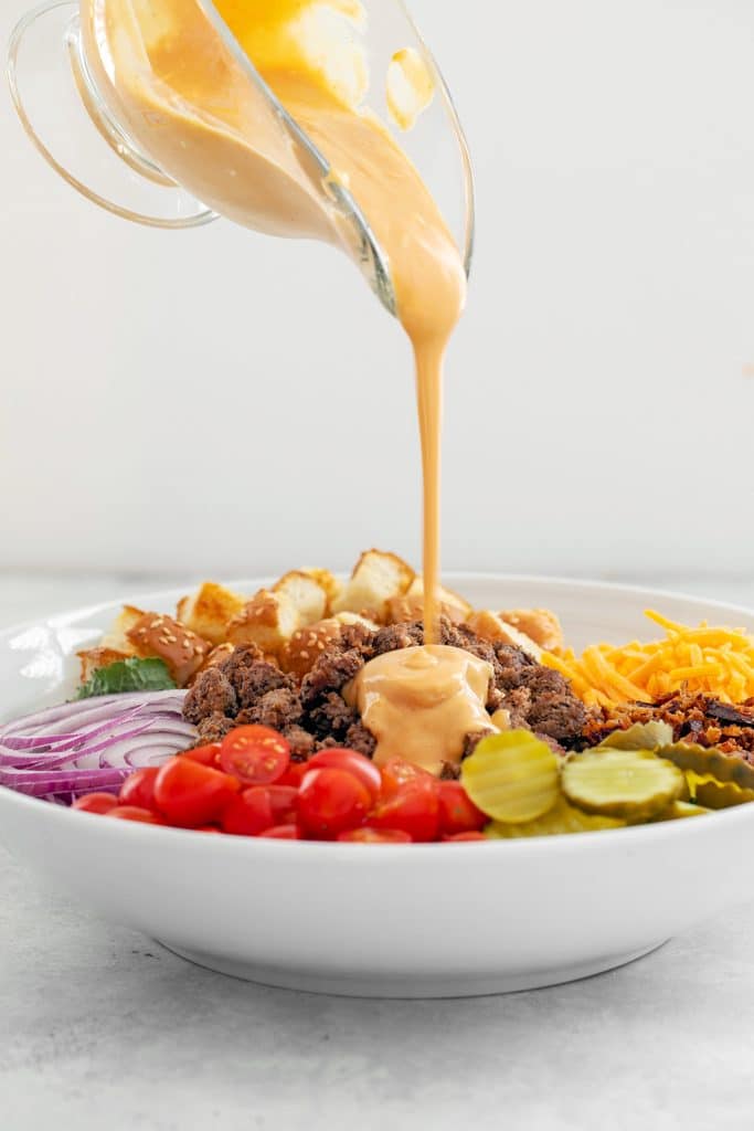 Head-on view of white serving bowl of bacon cheeseburger kale salad with gravy boat filled with "secret sauce" dressing being poured over the top