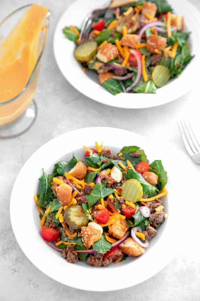 Bird's eye view of two white bowls of bacon cheeseburger kale salad, including baby kale, chopped tomatoes, sliced red onion, dill pickle rounds, shredded cheddar, crumbled bacon, hamburger bun croutons, and ground beef with gravy boat of dressing and fork in the background