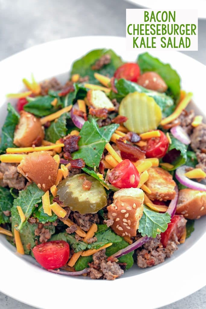 Overhead close-up view of bacon cheeseburger kale salad including baby kale, ground beef, shredded cheddar, dill pickle rounds, chopped tomatoes, red onion, crumbled bacon, and hamburger bun croutons in a white bowl with recipe title at top