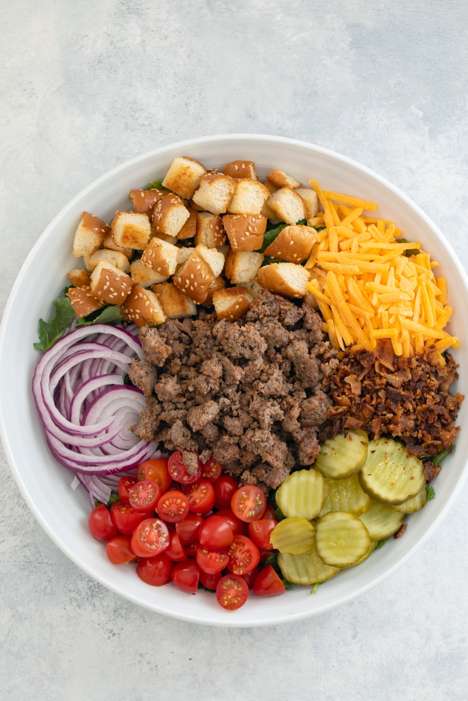 Overhead view of salad ingredients arranged in white serving bowl before tossing, including bed of baby kale with sliced red onion, chopped tomatoes, dill pickle rounds, crumbled bacon, shredded cheddar, hamburger bun croutons, and ground beef