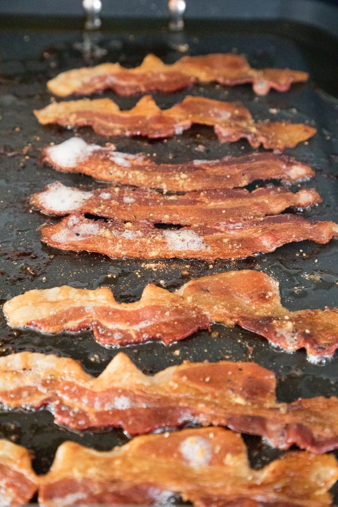 Bacon frying on a griddle with cracked black pepper and ground fenugreek