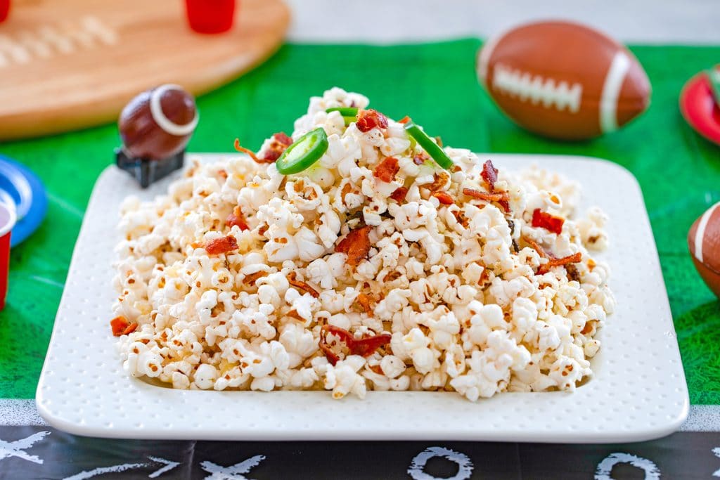 Landscape photo of bacon jalapeño cheddar popcorn on a white platter on a football field tablecloth with mini football and football cutting board in background