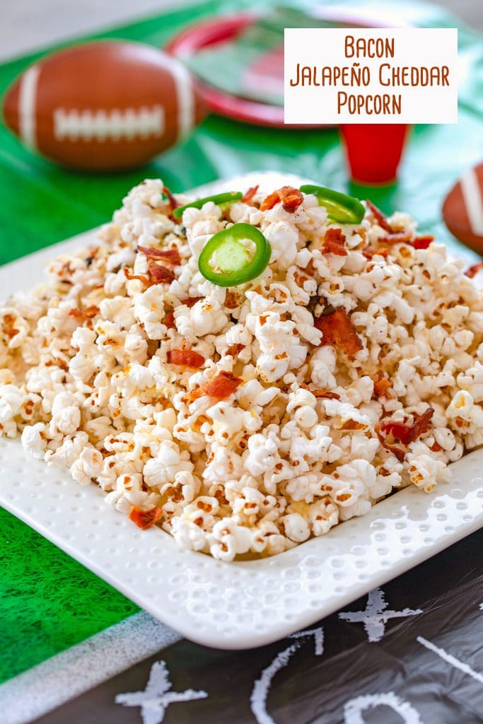 Head-on view of platter of bacon jalapeño cheddar popcorn on a football field tablecloth with plates, cups, and footballs in background and recipe title at top of image