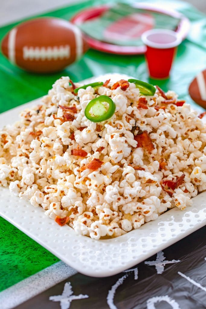 Head-on view of platter of bacon jalapeño cheddar popcorn on a football field tablecloth with plates, cups, and footballs in background and recipe title at top of image