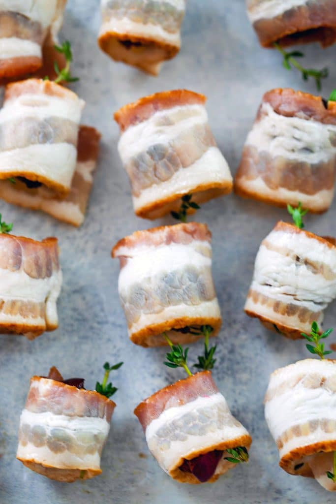 Several bacon wrapped cherries with thyme on a gray surface before being baked