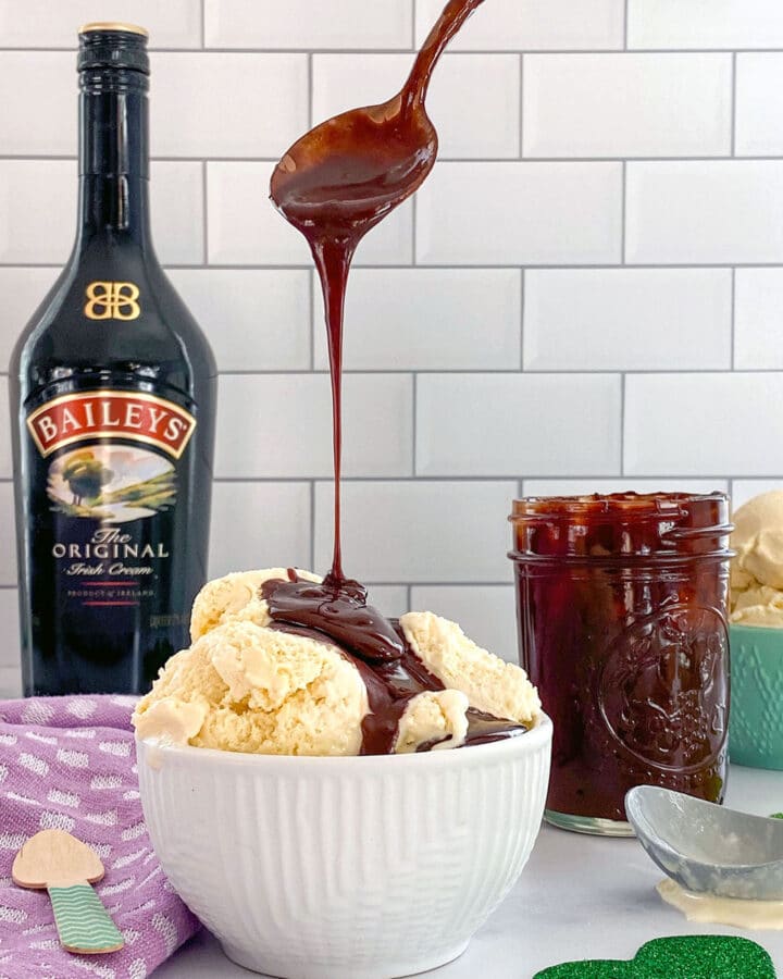 Head-on view of a bowl of Baileys ice cream with a spoon drizzling Baileys hot fudge sauce over it and a bottle of Baileys Irish Cream in background