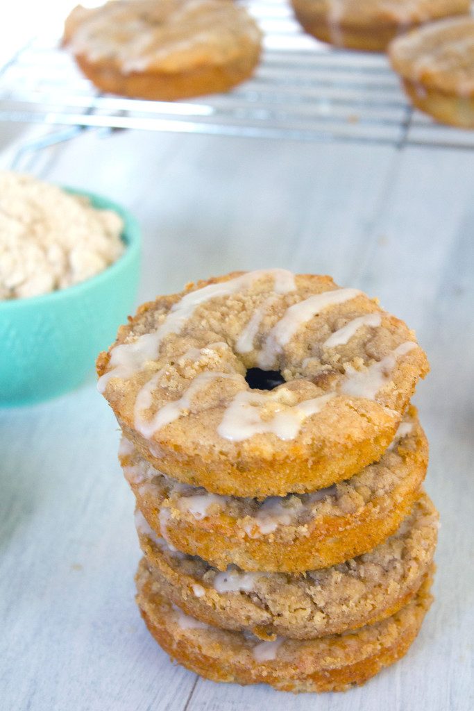 Head-on close-up view of four baked apple coffee cake doughnuts with bowl of streusel and more doughnuts cooling in background 