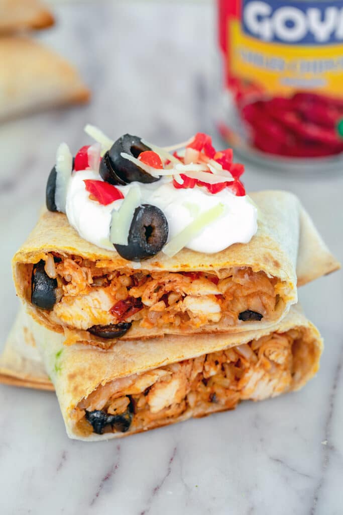 Head-on view of a baked chicken chimichanga cut in half on a marble surface and topped with sour cream, tomatoes, cheese, and olives, with can of chipotle peppers in background