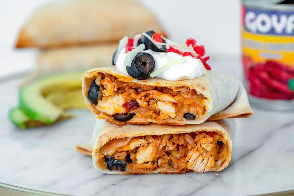 Landscape head-on view of baked chicken chimichanga cut in half and topped with sour cream, shredded cheese, olives, and tomatoes with sliced avocado, can of chipotle peppers, and more chimichangas in the background