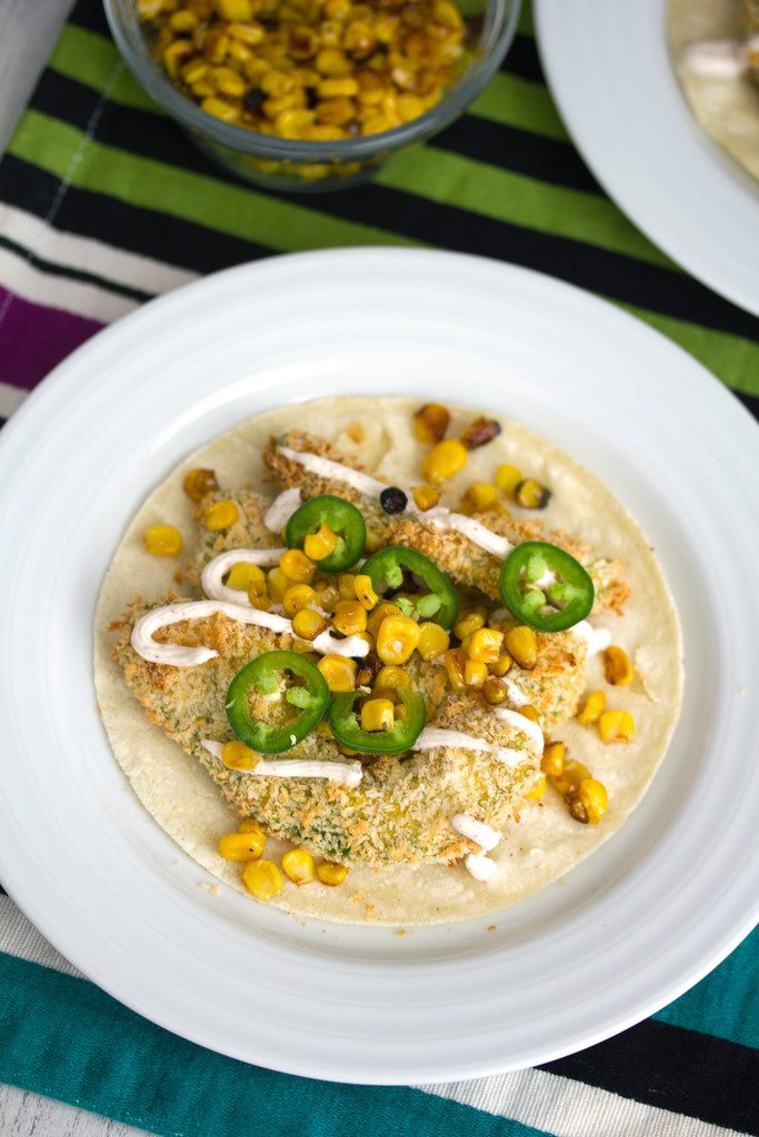 Overhead view of a baked avocado taco with jalapeño, grilled corn, and Greek yogurt sauce on a large white plate with bowl of grilled corn in the background