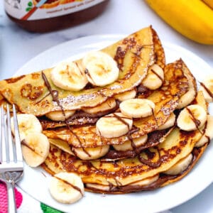 Three banana crepes on plate with Nutella drizzled on and sliced bananas on top