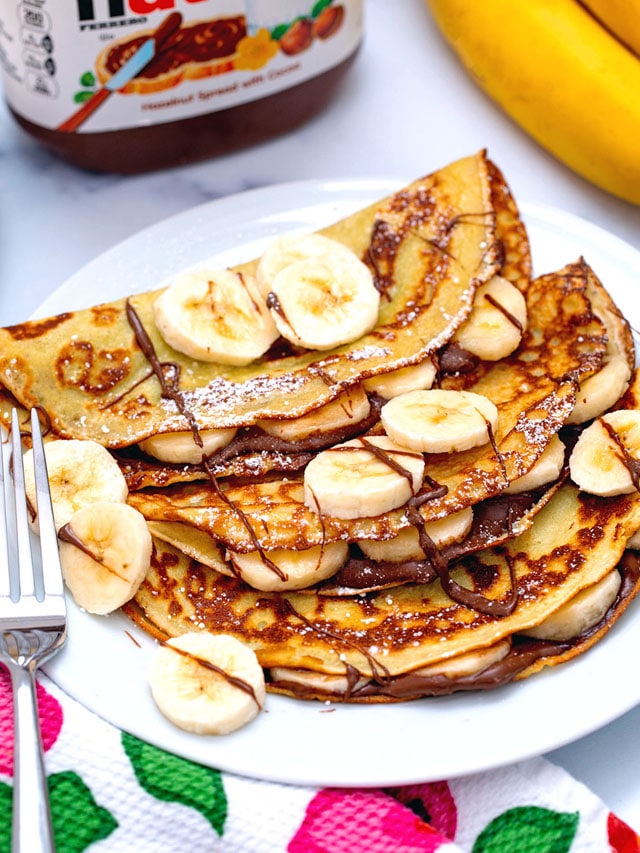 Overhead view of banana crepes topped with Nutella, sliced bananas, and confectioners' sugar with jar of Nutella in background