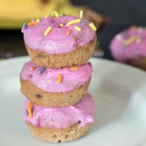 Closeup of three banana donuts for dogs with pink frosting and sprinkles stacked on a white plate with banana in background