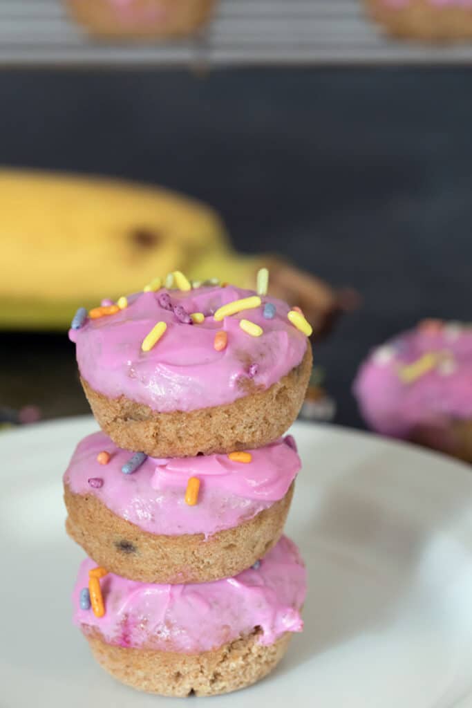 Stack of three donuts for dogs with pink icing and sprinkles on a white plate with banana in background