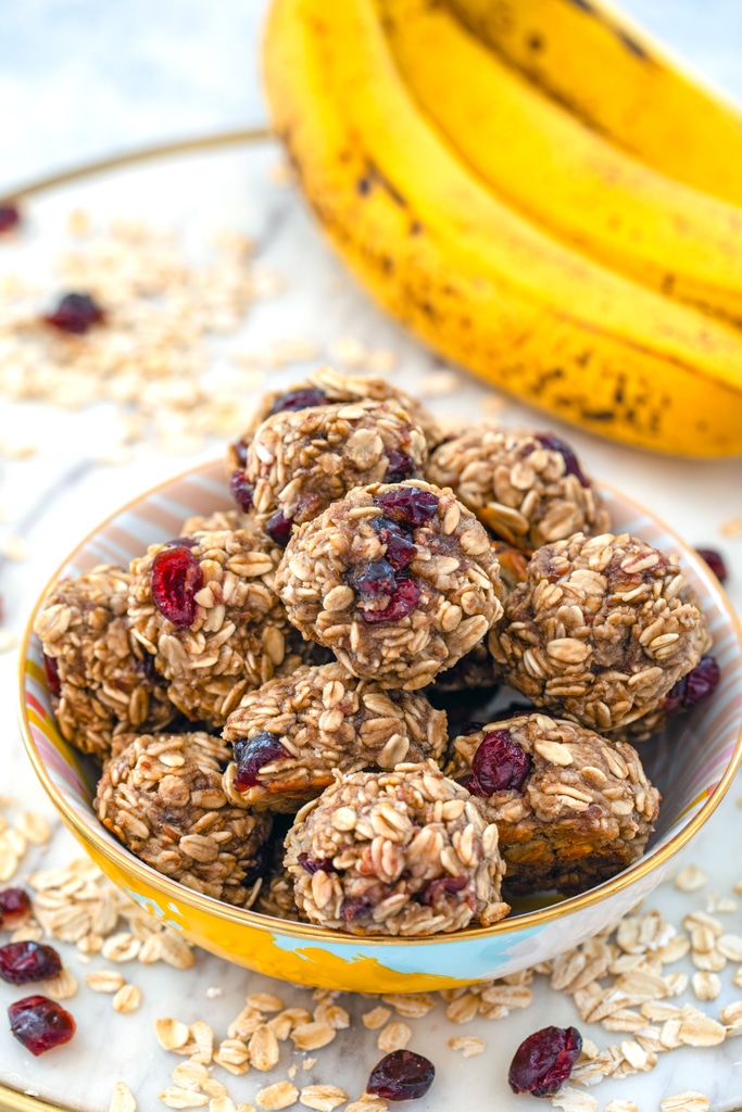 Head-on view of a bowl filled with banana maple cranberry cookies on a marble tray with oats and dried cranberries all around and bananas in background