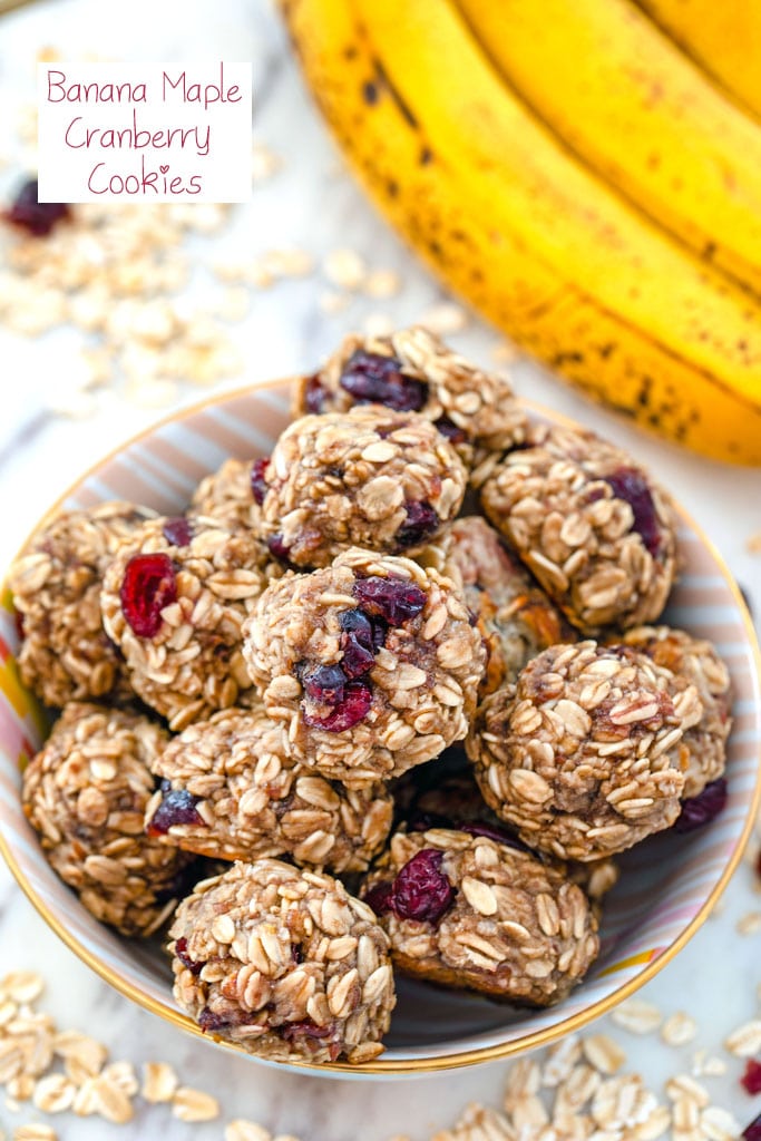 Overhead view of banana maple cranberry cookies in a bowl surrounded by oats and dried cranberries with bananas in background and recipe title at top