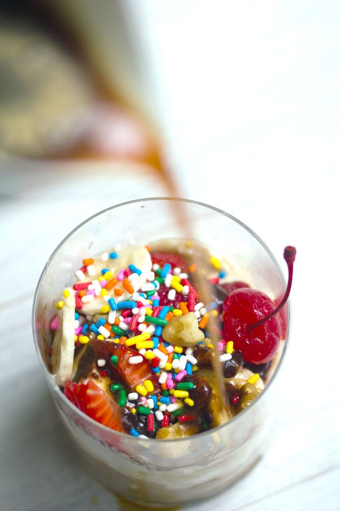 Birds eye view of cup of ice cream, bananas, strawberries, sprinkles, nuts, and a cherry, with espresso being poured over the top for banana split affogato