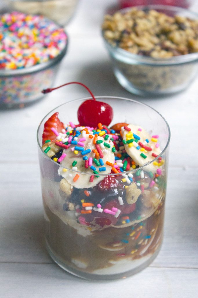 From above shot of banana split affogato with lots of sprinkles and a cherry on top and bowls of sprinkles and nuts in the background