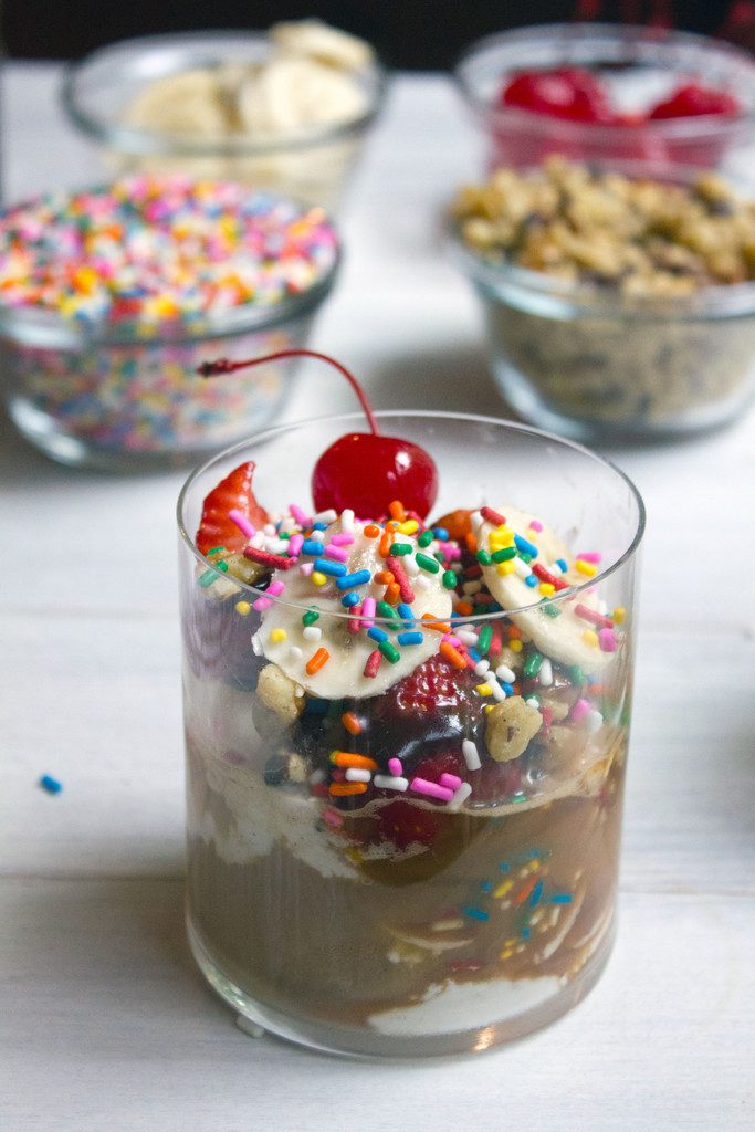 Close up of banana split affogato with a cherry on top and bowls of sprinkles, nuts, cherries, and sliced bananas in the background