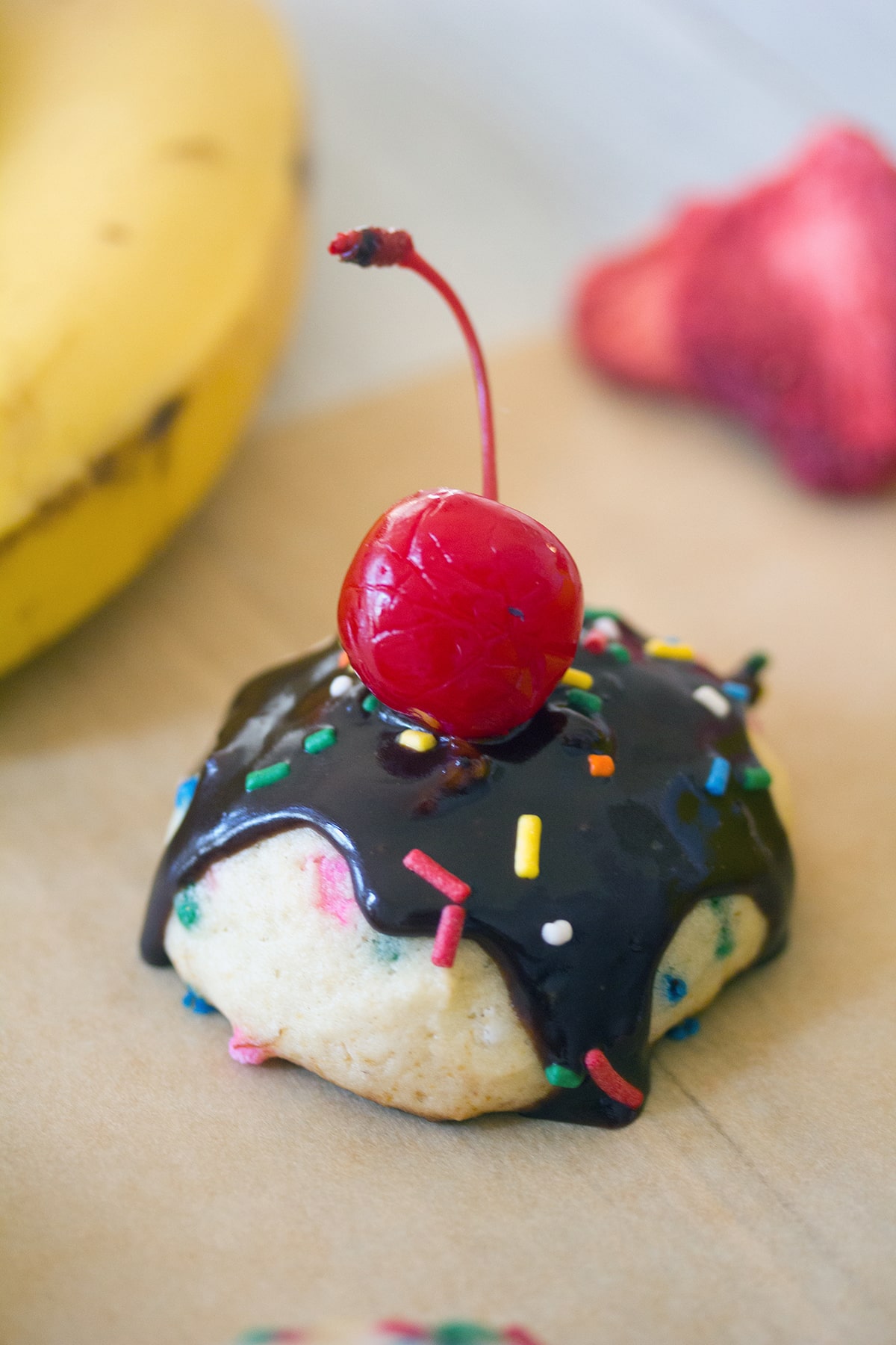 Overhead view of a banana split cookie topped with chocolate sauce, sprinkles, and a cherry with bananas and strawberries in the background.