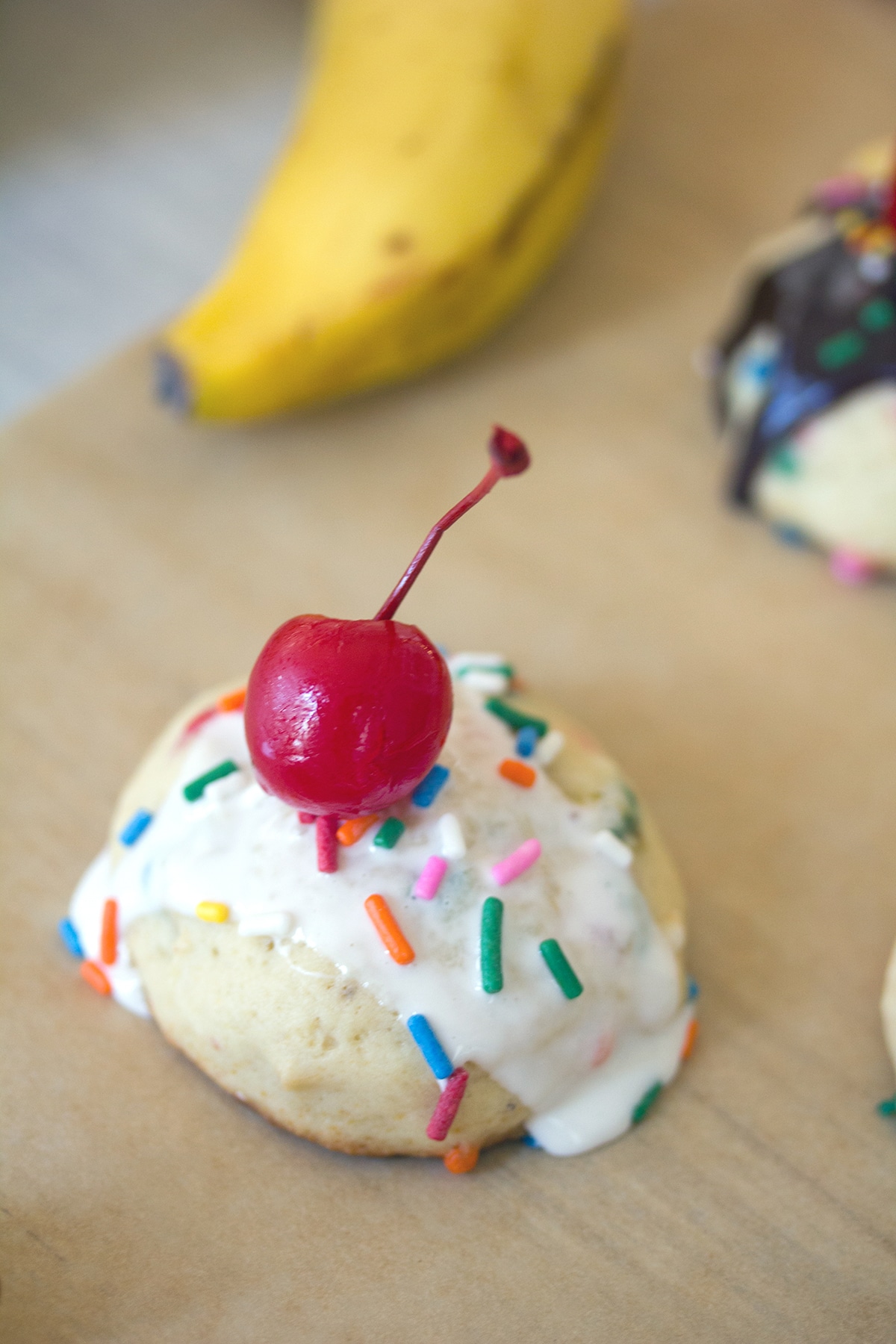 Overhead view of a banana split cookie topped with marshmallow sauce, sprinkles, and a cherry with bananas and more cookies in the background.
