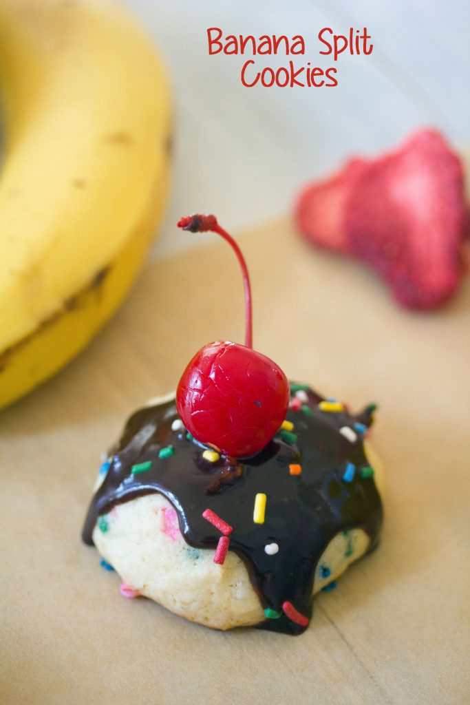 Head-on view of a banana split cookie topped with chocolate sauce, sprinkles, and a cherry with bananas and strawberries in the background and recipe title at top