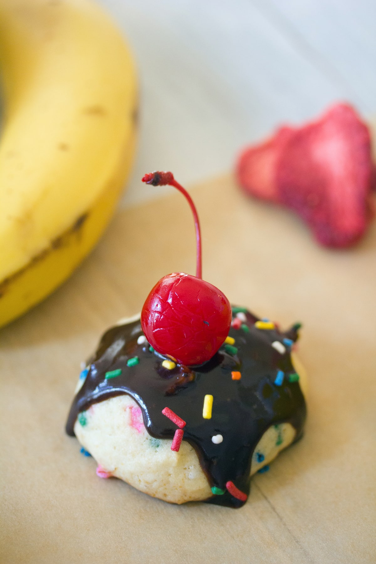 Overhead view of a banana split cookie topped with chocolate sauce, sprinkles, and a cherry with a banana and freeze-dried strawberries in the background.