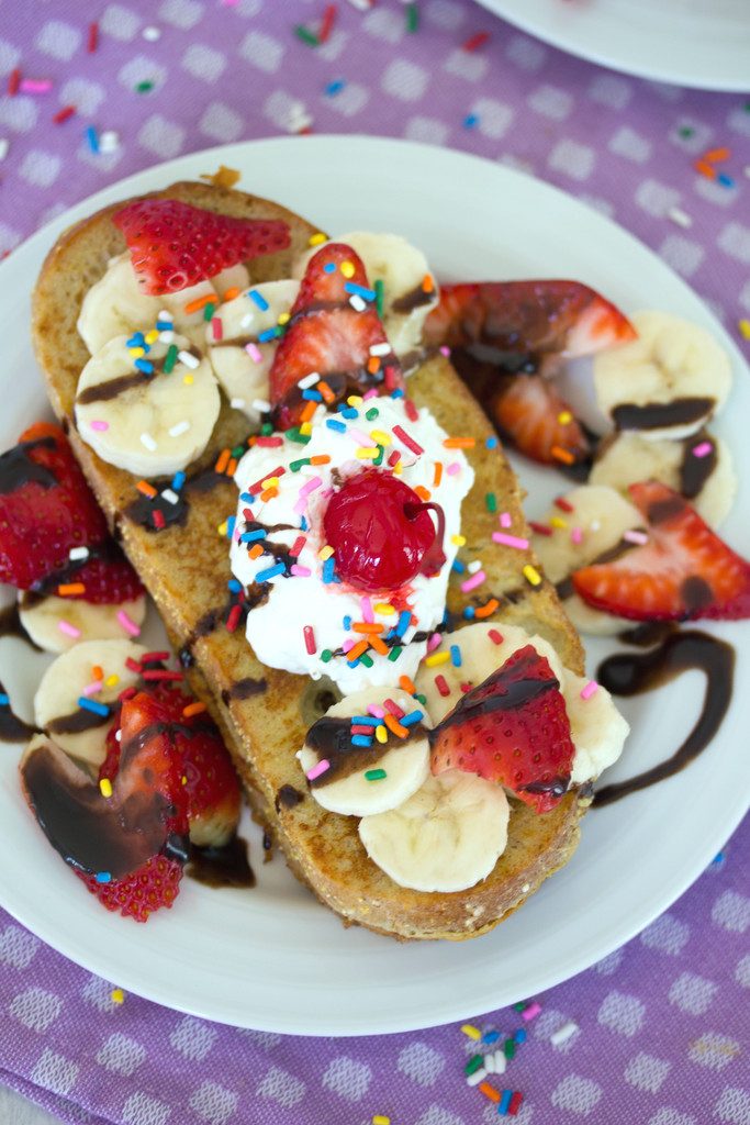 Overhead view of slice of banana split french toast topped with strawberries, bananas, chocolate, whipped cream, sprinkles, and a cherry