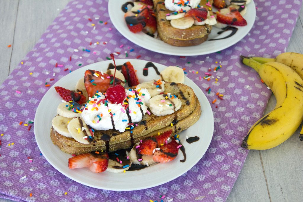 Landscape view of a plate of banana split french toast topped with bananas, strawberries, chocolate sauce, whipped cream, sprinkles, and a cherry with a second plate and whole bananas in the background