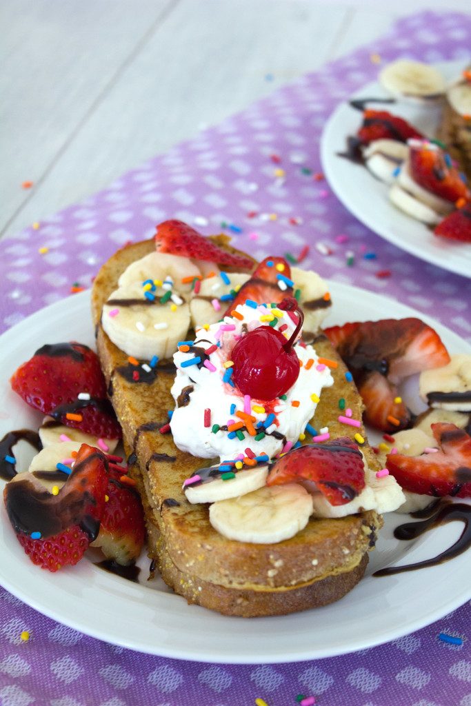 Head-on closeup image of a slice of banana split french toast topped with strawberries, bananas, chocolate sauce, whipped cream, sprinkles, and a cherry on a purple tea towel