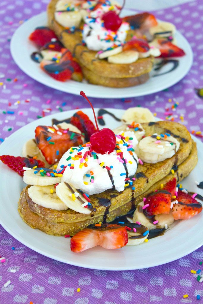 Head on view of a white plate with banana split french toast topped with bananas, strawberries, whipped cream, chocolate, sprinkles, and a cherry with a second plate in background