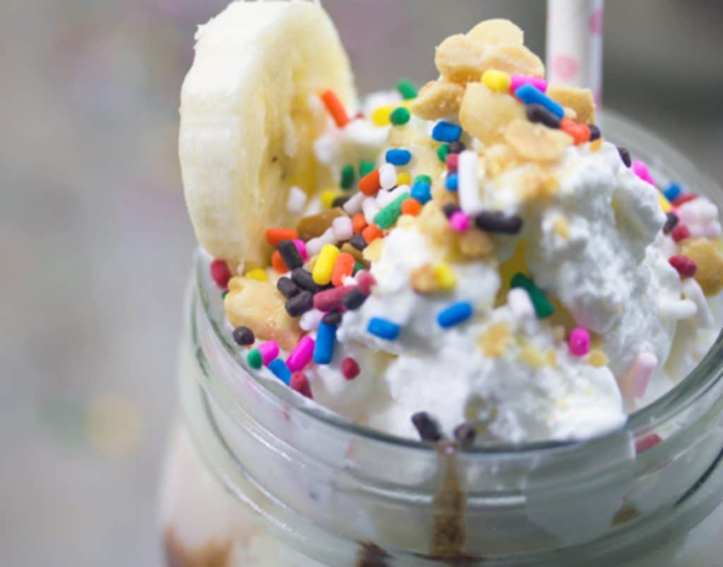 Closeup landscape view of the top of a banana split vodka milkshake, showing whipped cream, sprinkles, banana slice, and peanuts