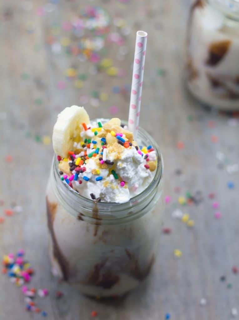 Centered image of banana split vodka milkshake with whipped cream, sprinkles, peanuts, banana slice, and straw, surrounded by sprinkles