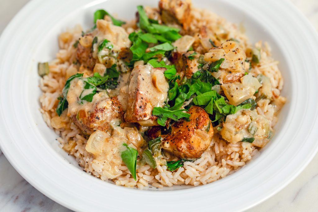 Landscape view of a white bowl filled with brown rice that's topped with basil chicken in coconut curry sauce with fresh basil on top