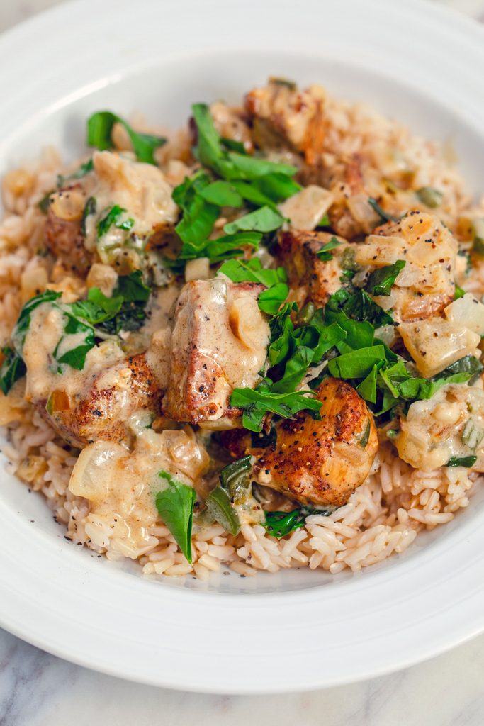 Overhead closeup view of a white bowl filled with brown rice and topped with basil chicken in coconut curry sauce with extra fresh basil on top