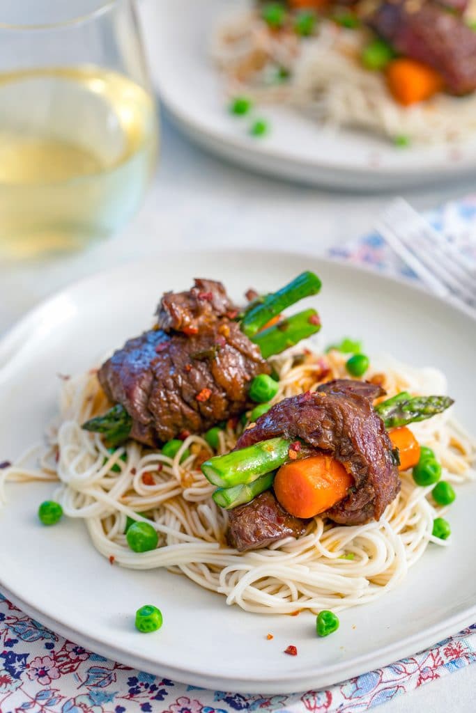 Head-on view of two steak roll-ups with asparagus and carrots over a bed of noodles and peas with a second plate and glass of white wine in the background