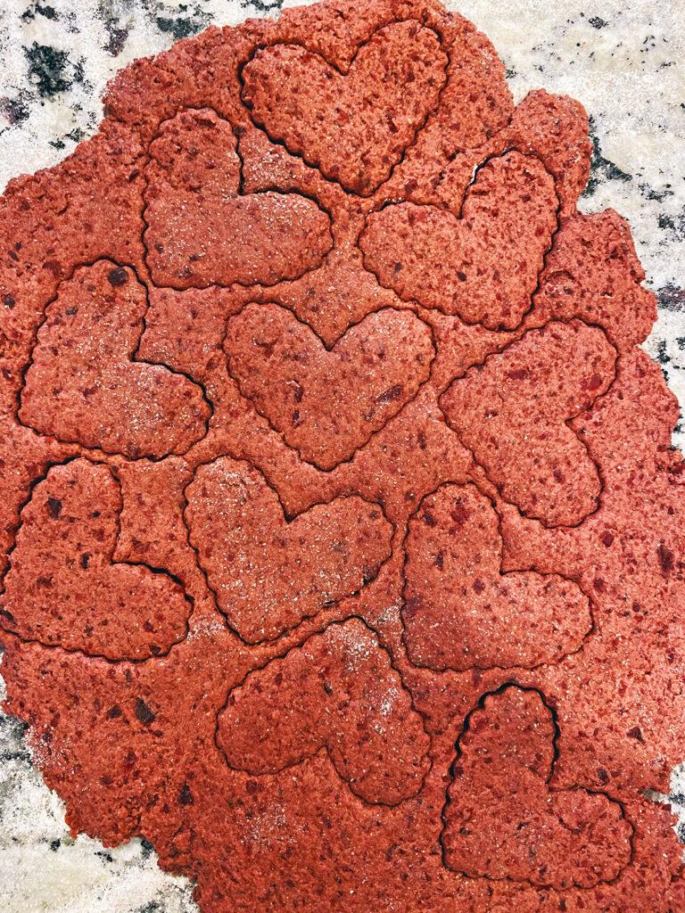 Heart shapes being cut out of beet cookie dough on counter