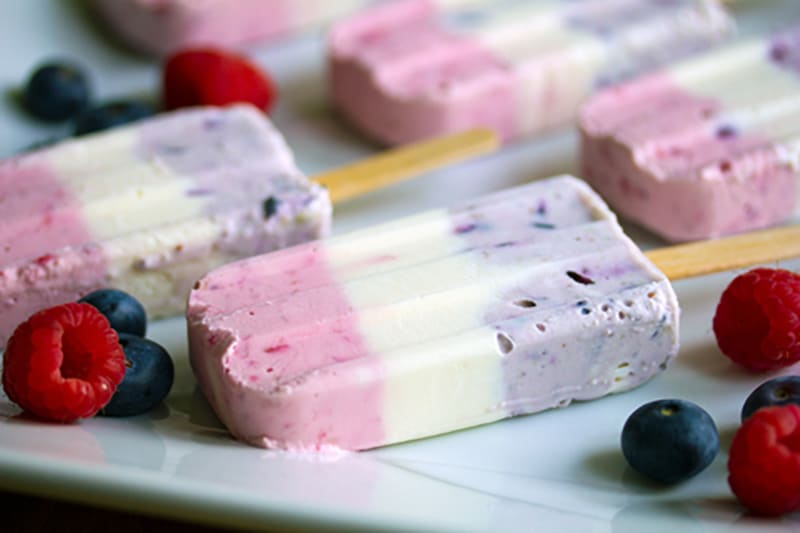 Head-on view of Berry Greek Cheesecake Popsicles showing red (pink-ish), white, and blue (purple-ish) layers, surrounded by raspberries and blueberries