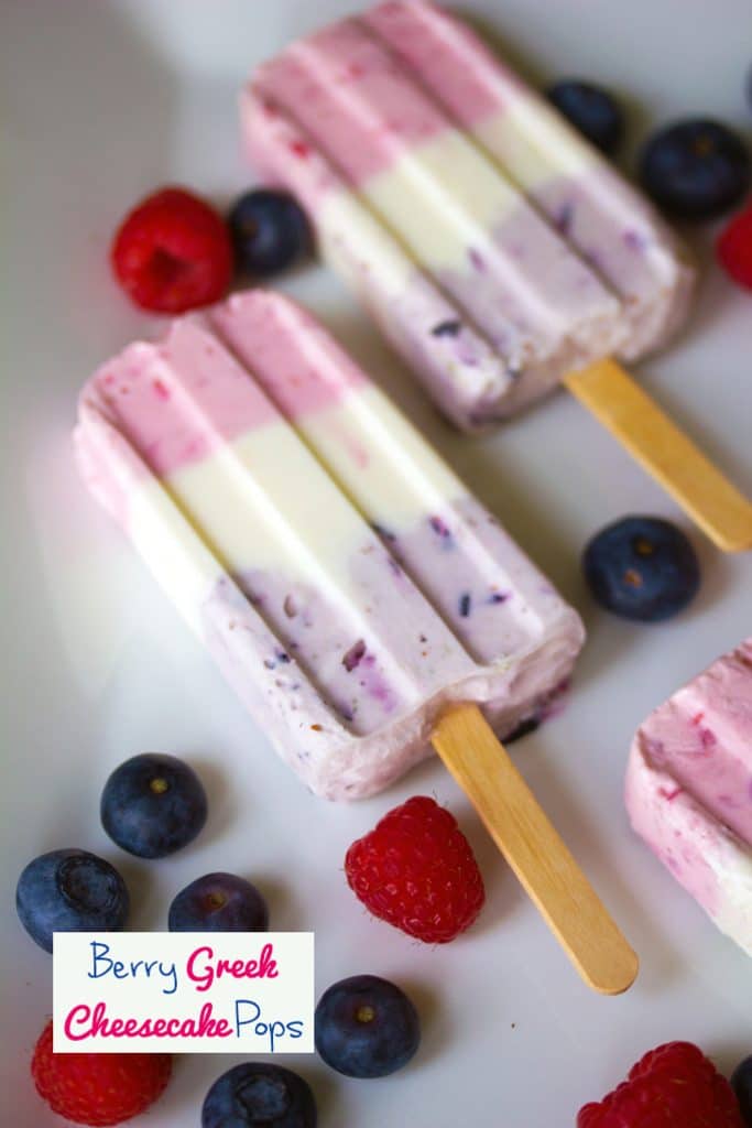 Overhead view of Berry Greek Yogurt Popsicles on a white plate surrounded by blueberries and raspberries and "Berry Greek Cheesecake Pops" text at the bottom