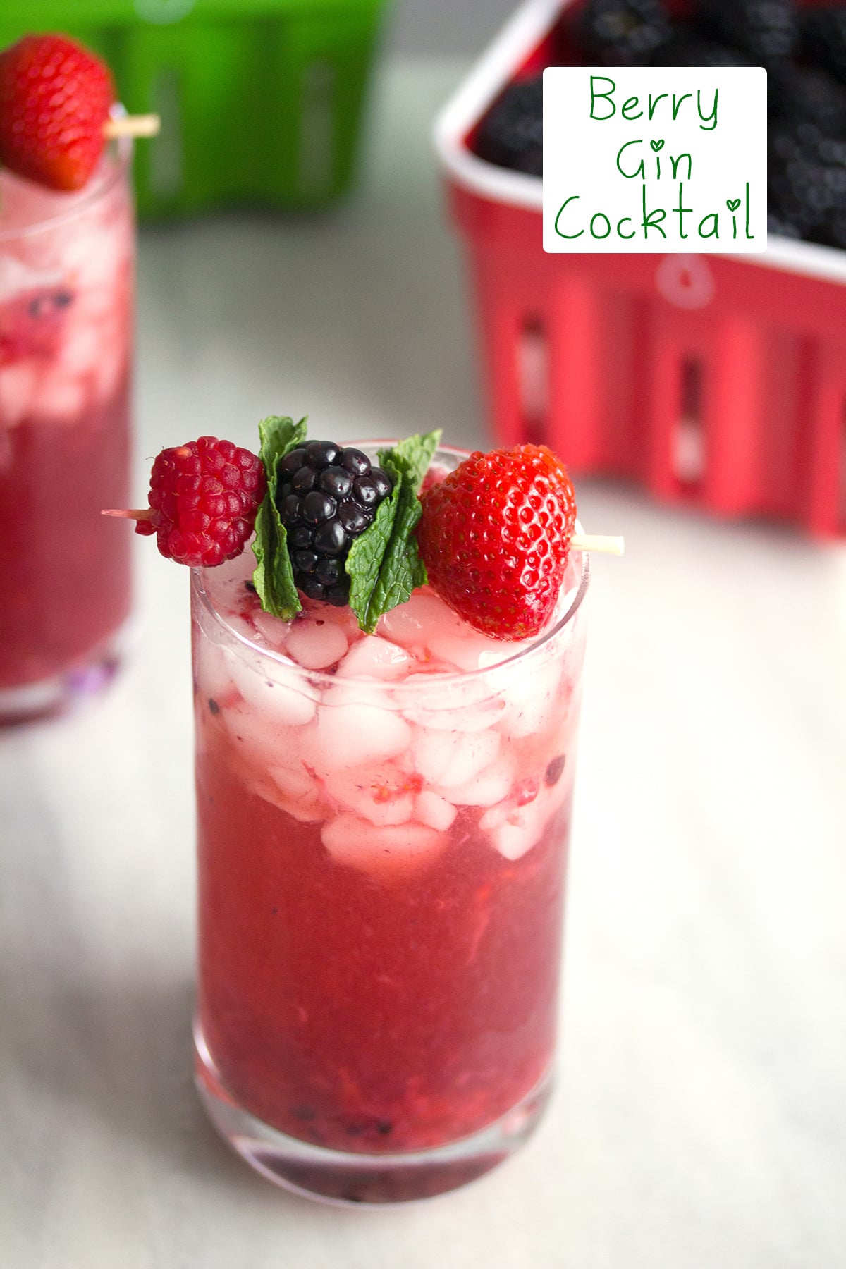 A red berry cocktail packed with ice and a berry and mint garnish with baskets of berries in the background and "Berry Mint Gin Smash" text at the top.