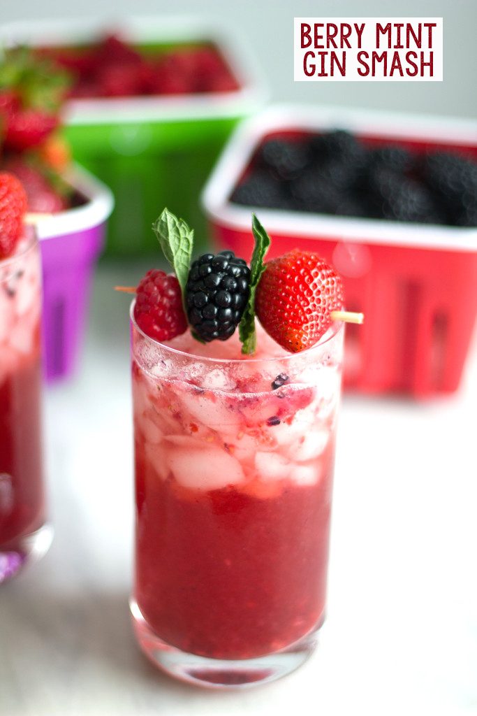 A red berry cocktail packed with ice and a berry and mint garnish with baskets of berries in the background and "Berry Mint Gin Smash" text at the top