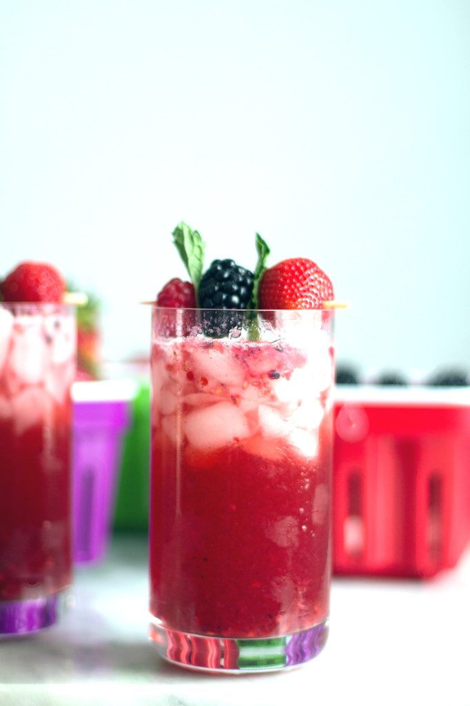 A head on view of a red berry cocktail with a garnish with raspberry, blackberry, and strawberry and mint leaves