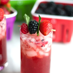 Berry mint gin smash cocktail with raspberry, blackberry, strawberry, and mint garnish with baskets of berries in background