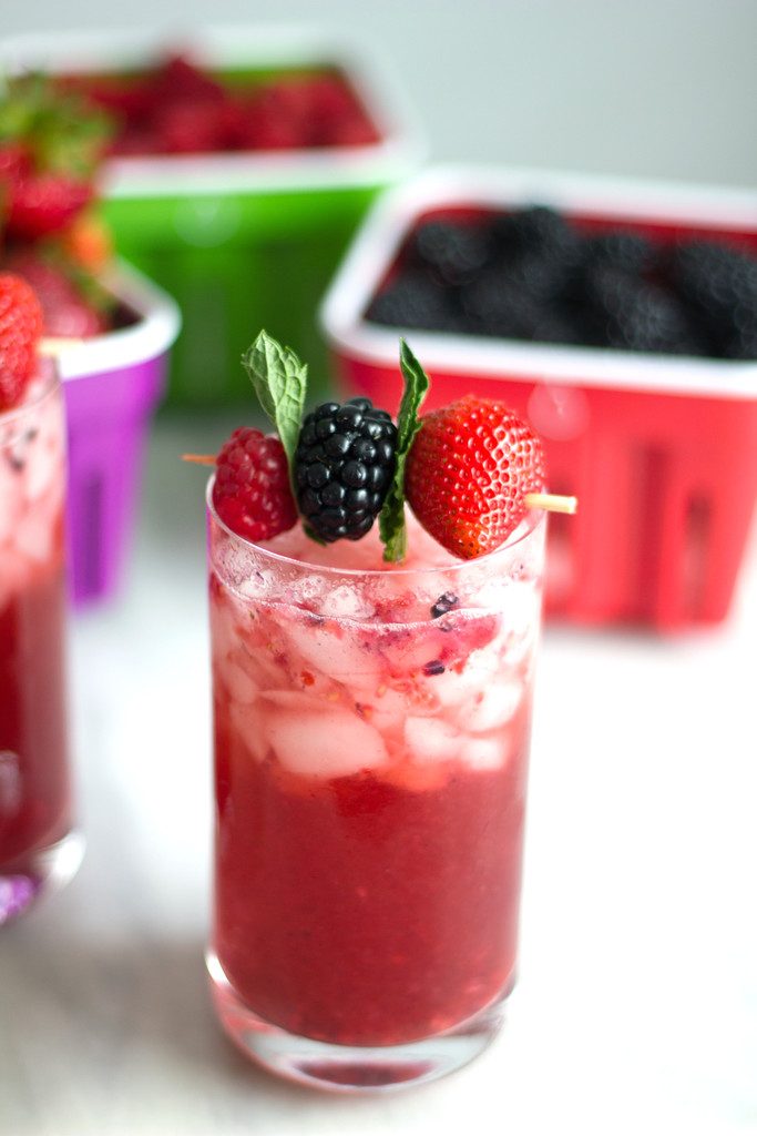 A head on view of a red cocktail with a berry and mint garnish with berry filled baskets in the background