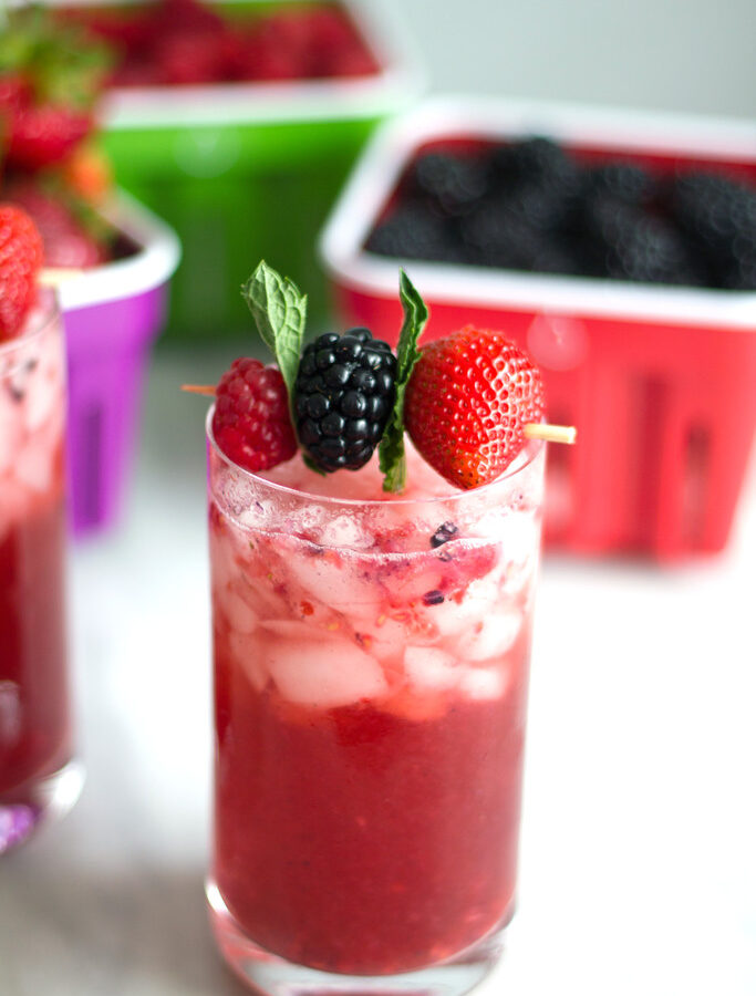 Berry mint gin smash cocktail with raspberry, blackberry, strawberry, and mint garnish with baskets of berries in background