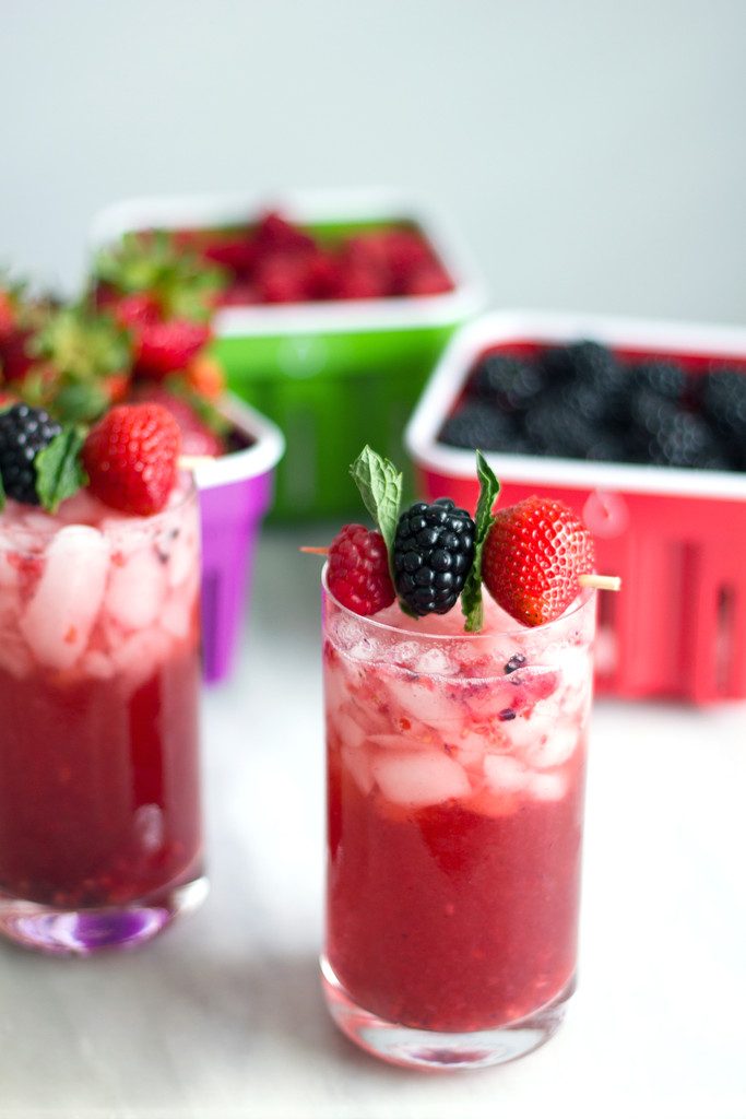 An arial view of two red berry cocktails with berry and mint garnishes and blackberries and raspberries in berry baskets in the background