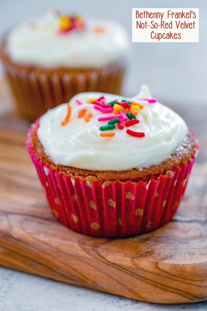 Head-on view of one of Bethenny Frankel's not-so-red velvet cupcakes with cream cheese frosting and rainbow sprinkles on a wooden board with a second cupcake in the background and recipe title at top