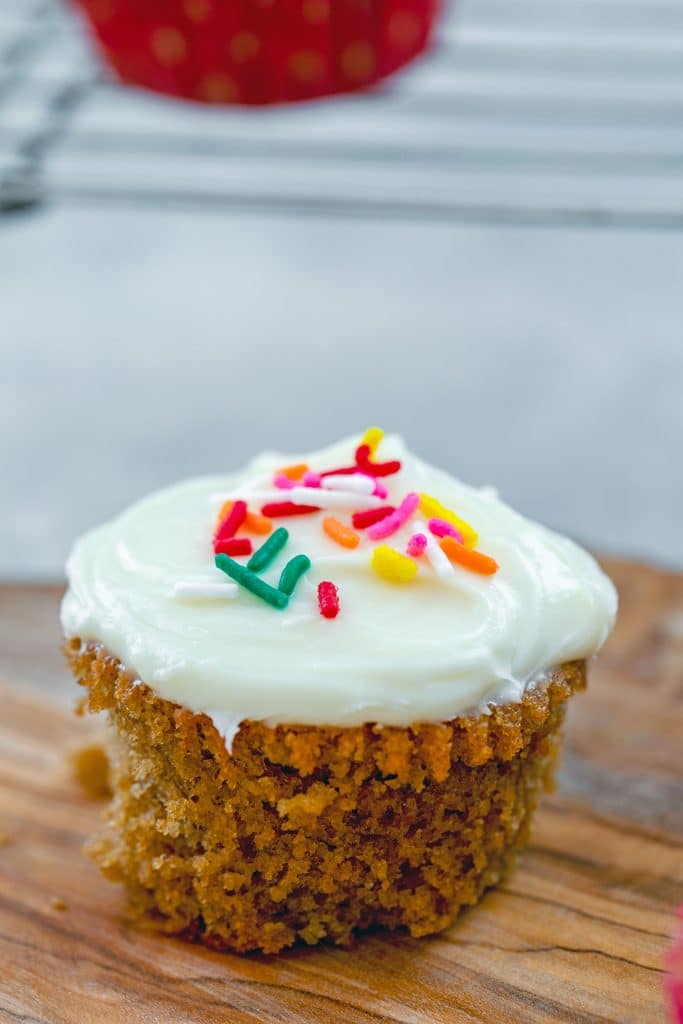 Head-on view of one of Bethenny Frankel's not-so-red velvet cupcakes with cream cheese frosting and rainbow sprinkles out of cupcake paper to show how delicate and crumbly they are sitting on wooden board