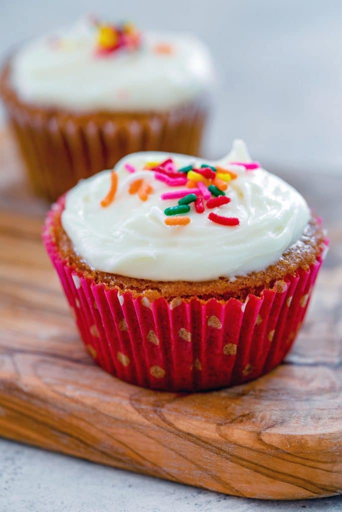 Head-on view of one of Bethenny Frankel's not-so-red velvet cupcakes with cream cheese frosting and rainbow sprinkles on a wooden board with a second cupcake in the background